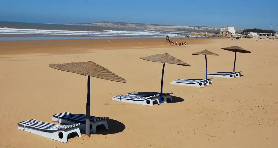 Sidi Kaouki beach with parasols, camels on the sand and the white marabout at the end of the bay